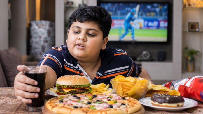 A realistic Indian school-age child (around 10–12 years old) sitting alone at a dining table filled with unhealthy fast foods such as pizza, burger, french fries, chips, sugary soft drinks, and packaged snacks. The child appears overweight and is reaching for a cold drink while looking tempted. In the background, a television is playing a sports match with blurred fast-food advertisements, symbolising the influence of marketing on children's eating habits. The room resembles a modern Indian middle-class home. The focus is on the child and the unhealthy food, with natural lighting, shallow depth of field, highly detailed, editorial health news photography style, ultra-realistic, 8K quality, cinematic composition, no text, no logo, no watermark, no infographic, no charts, no labels, no medical equipment, no exaggerated expressions.